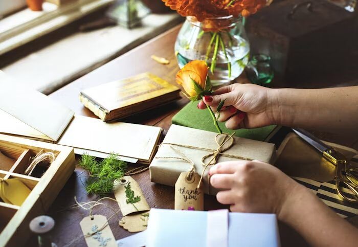 Tangan memegang bunga mawar oranye di atas kotak hadiah yang dikemas dengan kertas kraft dan tali.