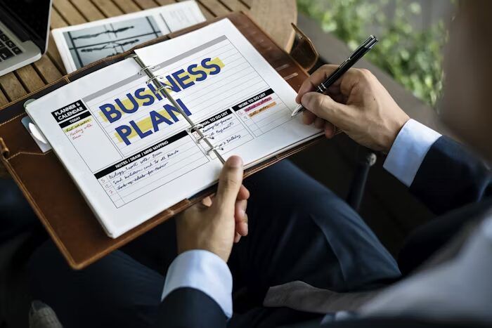 Tangan menulis pada lembaran bisnis plan di Uprint.id
