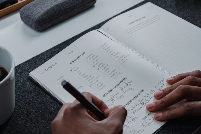 Tangan menulis dalam buku agenda dengan pena di meja kerja.