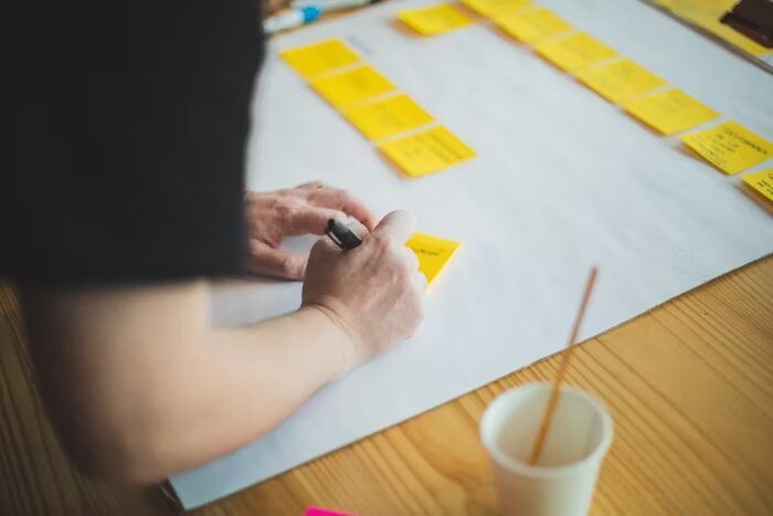 Tangan memegang marker dan menulis di sticky notes, ditempatkan di atas lembaran kertas.